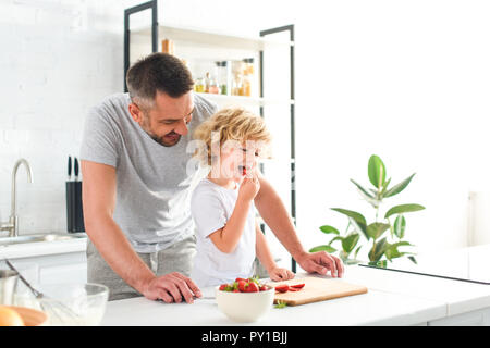 Lächelnd Vater steht in der Nähe von Son, während er Erdbeere in Küche essen Stockfoto