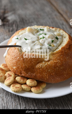 Vertikale Nahaufnahme von einem Brot Schüssel von New England Clam Chowder auf einem rustikalen Holztisch mit einem Löffel und Oyster Cracker. Stockfoto