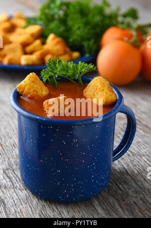 Nahaufnahme von einem blauen Becher mit frischen, hausgemachten Tomatensuppe gefüllt. Tomaten, Petersilie und Croutons unscharf im Hintergrund. Stockfoto