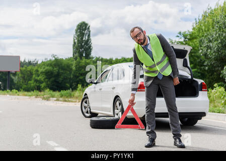 Gut aussehender Geschäftsmann in Licht grüne Weste, STOP-Schild auf der Straße in der Nähe von Broken Auto Stockfoto