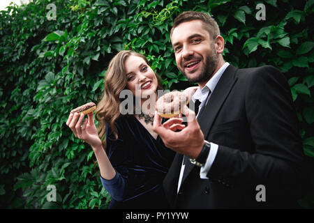 Portrait von lächelnden modischen Paar mit Schokolade Donuts mit grünem Laub hinter Stockfoto