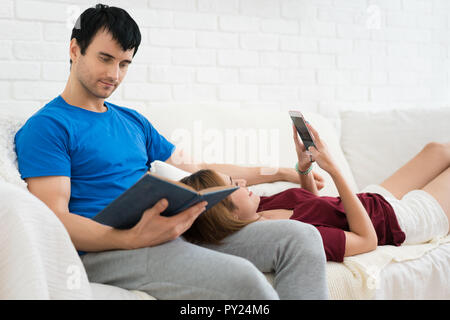 Junges Paar auf dem Sofa entspannen in Ihrem Wohnzimmer zu Hause. Mit Smartphone und ein Buch lesen. Stockfoto