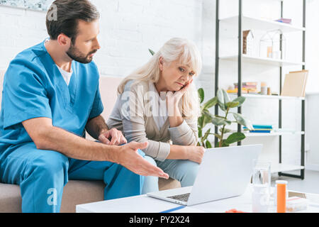 Verärgert senior Frau am Laptop auf der Suche beim Sitzen auf der Couch mit Sozialarbeiterin Stockfoto