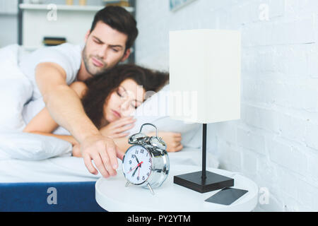 Junger Mann erreichen für Wecker im Schlaf mit Freundin im Bett Stockfoto