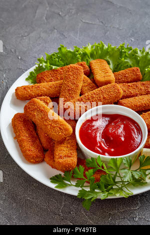 Panierte Fischstäbchen auf eine weiße Platte mit Zitrone, Salatblätter und Tomatensauce serviert, vertikale Ansicht von oben Stockfoto