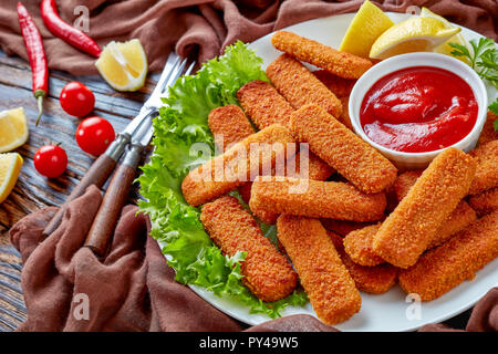 Panierte Fischstäbchen auf eine weiße Platte mit Zitrone, Salatblätter und Tomatensauce serviert, horizontale Ansicht von oben, close-up Stockfoto