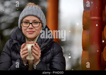 Schöne junge Dame in warme Kleidung und lächelnd in die Kamera halten Tasse heißes Getränk und in der Nähe von Haus auf dem Land auf wunderbare Win Stockfoto