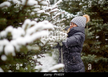 Seitenansicht der hübsche junge Frau in warme Kleidung lächelnd und berühren Nadelbaum Baum mit Schnee auf erstaunliche Winter Tag abgedeckt Stockfoto