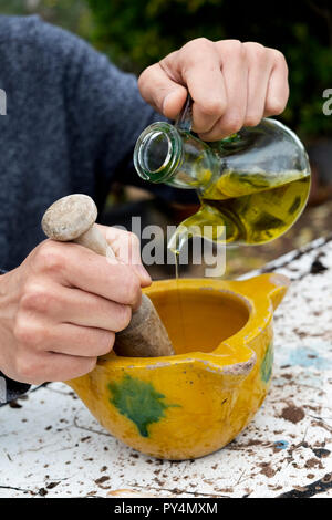 Nahaufnahme eines jungen kaukasischen Mann draußen etwas Aioli mit einer hölzernen Mörser in einem gelben Mörser aus Keramik, durch Zugabe von Olivenöl aus einem Glas Cruet Stockfoto
