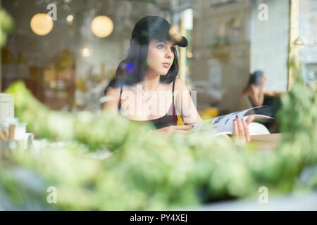 Portrait einer jungen schönen Mädchen allein in einem Cafe sitzen und Lesen einer Modezeitschrift. Durch das Fenster geschossen. Stockfoto