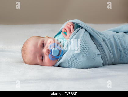 Baby Festlegung in eine blaue Decke mit Blick auf die Kamera mit verschlafenen Augen und Schnuller im Mund gewickelt. Stockfoto