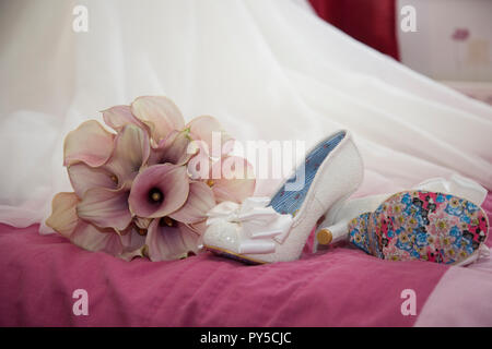 Bräute Schuhe auf dem Bett neben ihr Bouquet von rosa Calla Lilien sitzt am Ende ihrer Hochzeit Kleid Zug wartet an Ihrem Hochzeitstag getragen zu werden. Stockfoto