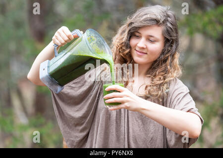 Junge Frau, Draußen, Draußen, holding Kunststoff mixer Behälter, Gießen grünes Gemüse, kale Smoothie in Glas Stockfoto