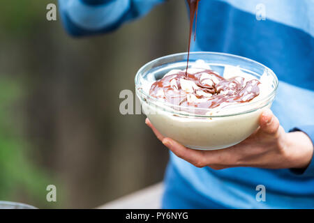 Nahaufnahme der junge Mann draußen, im Freien, holding Glas Schale hausgemachter, Raw vegan Vanille Eis, Schokolade Flüssigkeit hinzufügen Sauce vom Löffel Stockfoto