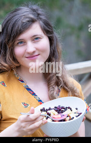 Nahaufnahme der jungen Frau, die weiblichen sitzen draußen auf Holz- Heim, Haus deck Holding, essen raw vegan Obstsalat aus Heidelbeeren, Bananen, Äpfel, blueber Stockfoto