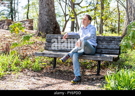 Glücklicher Mann im Kleid Shirt und Jeans sitzen entspannt auf einer Holzbank im sonnigen Park Wald im Frühling in Roanoke, Virginia Stockfoto