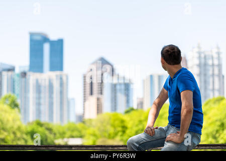 Junge Mann sitzt auf Geländer in Piedmont Park in Atlanta, Georgia rückblickend auf das malerische Stadtbild Skyline der Stadt Wolkenkratzer in der Innenstadt Stockfoto