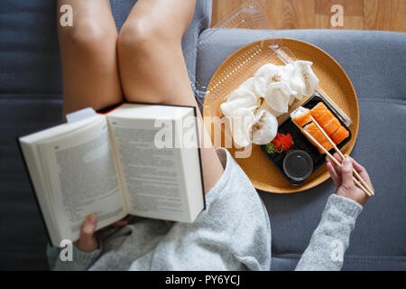 Junge Frau mit Buch und essen Sushi auf dem Sofa zu Hause. Ansicht von oben Stockfoto