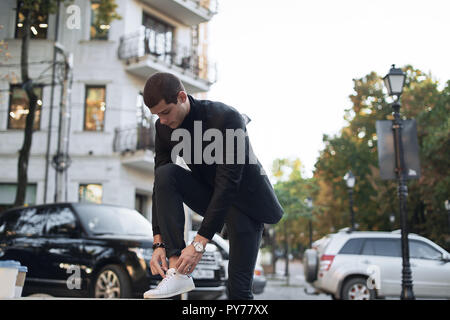 Attraktiver Mann auf der Straße zu binden Schnürsenkel auf seinem weißen Turnschuhe. Junge modische Geschäftsmann. Stockfoto