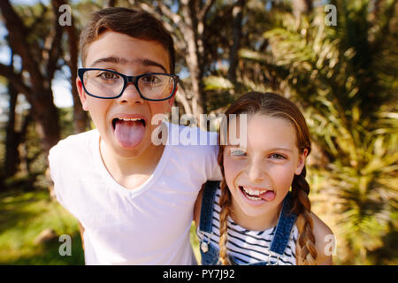 Fröhlicher Junge und Mädchen stehen in einem Park und Gesichter machen mit ihrer Zunge heraus. Nahaufnahme der lächelnden Kinder in einem Park stehen gemeinsam Spaß zu haben. Stockfoto