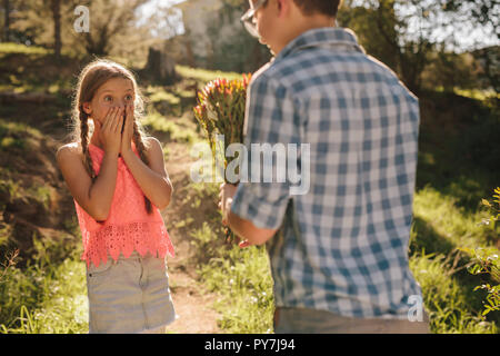 Junge schlägt ein Mädchen mit einem Blumenstrauß in einem Park stehen. Überrascht Mädchen auf den Mund mit Blick auf die Blumen in der Stockfoto