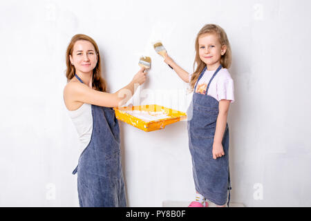 Reparatur in der Wohnung. Glückliche Familie, Mutter und Tochter in blauen Schürzen malt die Wand mit weißer Farbe Stockfoto