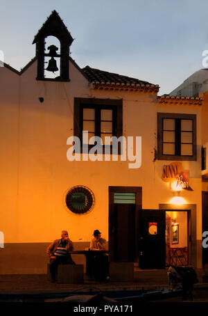 Zwei reife Männer sitzen am frühen Abend vor der Sunny Bar in Funchal, Madeira, während das Licht verblasst und die Straßenlaternen aufleuchten. Über ihnen ist ein kleiner Glockenturm Stockfoto