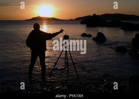 Selbstporträt des Fotografen, der ein Foto von Little Ross Island und dem Leuchtturm von Torrs Point über Kirkcudbright Bay, Dumfries und Galloway, Schottland, macht. Seine DSLR befindet sich auf einem Stativ und er verwendet eine Kabelfreigabe, um ein friedliches Sonnenuntergangsbild zu machen Stockfoto