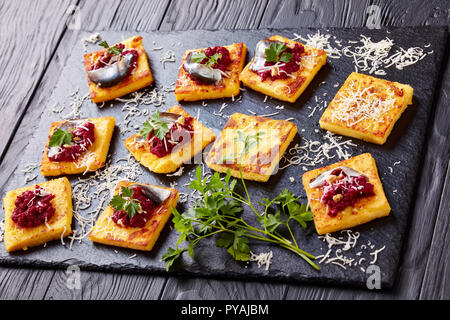 Close-up von Teilen der Quadrate mit cremiger Polenta rote Beete Pastete, gekrönt mit Sardellen und Petersilie auf einen schwarzen Stein Fach auf einer hölzernen Tisch serviert, v Stockfoto