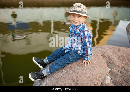 Ein kleiner Junge sitzt auf dem Rock bei einem Spaziergang im Park vor dem Hintergrund der See und eine schwimmende Ente. Stockfoto