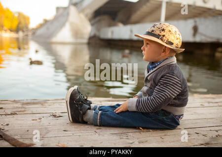 Ein kleiner Junge sitzt neben den See bei einem Spaziergang im Park vor dem Hintergrund der gelbe Bäume im Herbst und schwimmenden Enten. Stockfoto