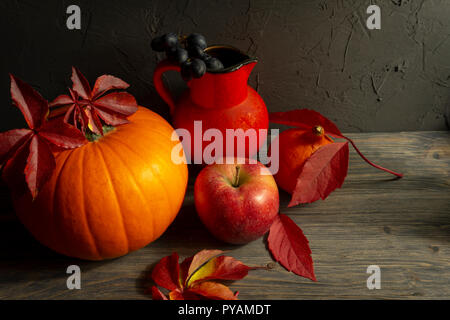 Zwei Kürbisse mit hellen Blätter im Herbst apple, blaue Trauben acorn und roten Kanne auf Holz- rustikalen Tisch gegen einen schwarzen rauen Beton grunge Wand. Herbst Stockfoto