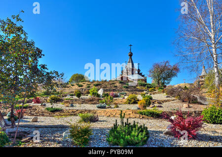 Zwerg Bäume, Büsche und Blumen auf dem steinigen Terrassen befestigte von gabionen am Hang - Vorgarten der orthodoxen Tempel. Kuppeln und Kreuze der Kirche Stockfoto