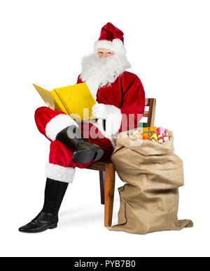 Traditionelle rot weiß Weihnachtsmann lesen Goldenes Buch mit Geschenk Beutel auf den Stuhl, der auf weisse Weihnachten Hintergrund isoliert Stockfoto