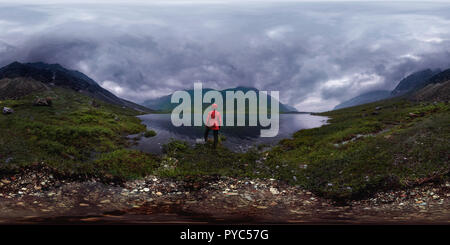 Man steht am See in roter Jacke bei bewölktem Himmel. Sphärisches Panorama 360 vr Stockfoto