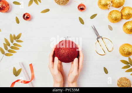 Weihnachten Dekorationen auf weißen Tisch Ansicht von oben. Women's Hände, die glänzend roten Ball baum Spielzeug unter den festlichen Golden flatlay Komposition. Stockfoto