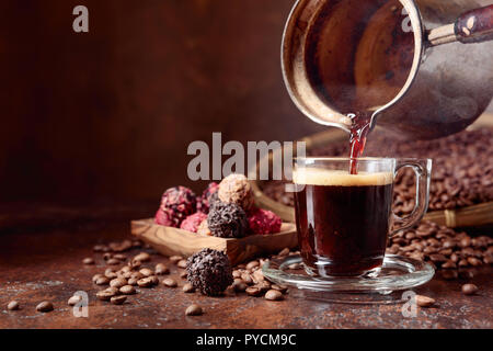Schwarzen Kaffee in ein kleines Glas Schale von einer Kupfer Kaffeemaschine gegossen. Kaffeebohnen und Pralinen auf einem alten Tisch. Kopieren Sie Platz. Stockfoto