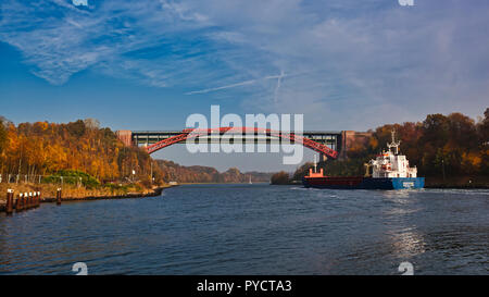 Nord-Ostsee-Kanal, alte Levensauer Hochbrücke', Kiel. Deutschland. Deutschland. Ein Container schiff nähert sich der Alten Brücke. Es ist Herbst und die Bäume an den Ufern des Kanals sind die Farben des Herbstes, reich an Rot-, Braun- und Gelbtönen. Diese reichen Farben sind, die von der Sonne auf der linken Kanalufer gebracht und das Wasser ist voll von Reflexionen. Ein Zug über die Brücke überquert. Stockfoto