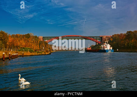 Nord-Ostsee-Kanal, alte Levensauer Hochbrücke', Kiel. Deutschland. Deutschland. Ein Container schiff nähert sich der Alten Brücke. Es ist Herbst und die Bäume an den Ufern des Kanals sind die Farben des Herbstes, reich an Rot-, Braun- und Gelbtönen. Diese reichen Farben sind, die von der Sonne auf der linken Kanalufer gebracht und das Wasser ist voll von Reflexionen. Ein Zug über die Brücke überquert und zwei Schwäne schwimmen auf dem Wasser im Vordergrund. Stockfoto