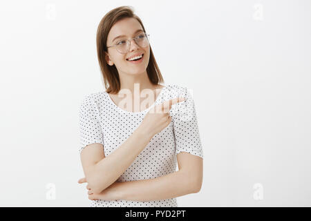 Taille-up Shot zufrieden und fröhlich gut aussehende Frau in Gläser, breit grinsend, gucken und nach rechts mit Zeigefinger, Fragen stellen über Kuriose, stehend über graue Wand Stockfoto