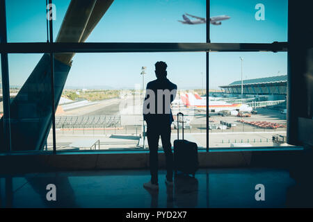 Ein junger und attraktiver Geschäftsmann sucht durch das Fenster in der Flughafen Terminal. Ein Flugzeug fliegt, während andere auf dem Grundstück geparkt werden Stockfoto