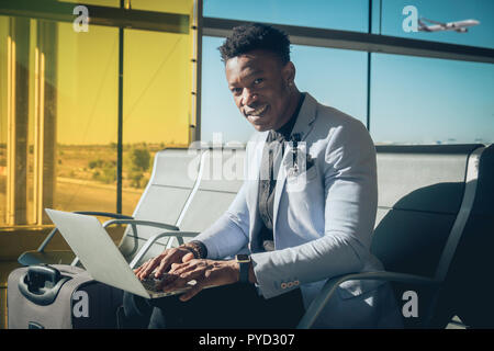 Eine junge und attraktive schwarze Unternehmer ist im Flughafen Terminal sitzt die Arbeit mit einem Laptop, und lächelnd. Er trägt einen Koffer und Tragekomfort Stockfoto