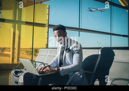 Eine junge und attraktive schwarze Unternehmer ist im Flughafen Terminal sitzt die Arbeit mit einem Laptop, und lächelnd. Er trägt einen Koffer und Tragekomfort Stockfoto