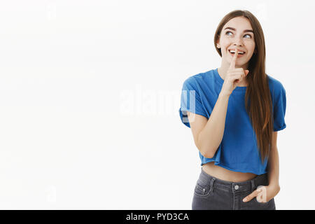 Verträumt und amüsiert charmante Freundin im blauen T-Shirt Blick auf die obere linke Ecke aufgeregt und lächelte breit, psst Geste mit dem Zeigefinger über den Mund halten Geheimnis oder lassen Sie sich überraschen Stockfoto