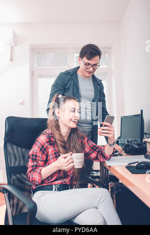 Lächelnd männliche und weibliche Mitarbeiter Spaß an der Arbeit brechen zusammen mit Smartphone, junge Frau, die Einstellungen auf Mobile für die gemeinsame Bild wi Stockfoto