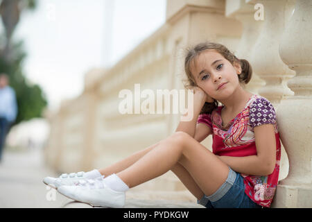 Adorable kleine Mädchen gekämmt mit Zöpfen draußen sitzen auf städtischen Boden. Stockfoto