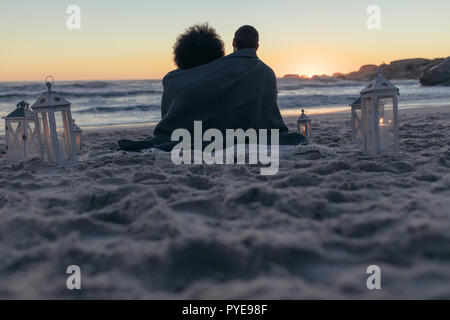 Ansicht der Rückseite des Paar am Strand zu sitzen eingehüllt in eine Decke den Sonnenuntergang. Paar sucht den Sonnenuntergang vom Strand. Stockfoto
