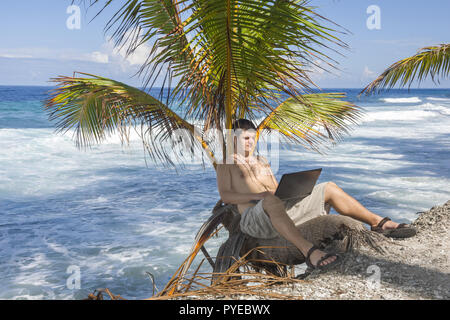 Mann bei der Arbeit am Strand. Attraktiver Mann mit Laptop in einer Palme sitzt an einem tropischen Strand. Junger Mann Arbeiten am Laptop auf der tropischen b Stockfoto
