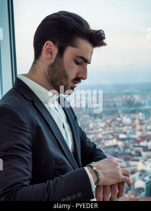 Portrait von stilvollen jungen Mann mit Anzug, stehend in der modernen Stadt, auf der Suche nach Armbanduhr die Zeit zu prüfen. Stockfoto