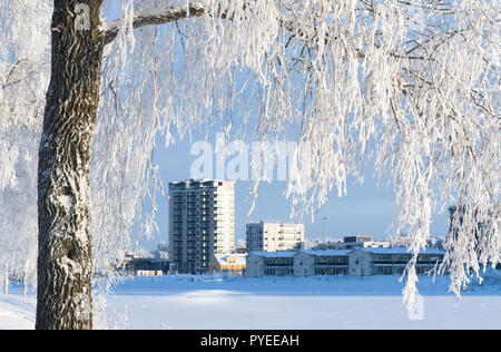 Winterlandschaft mit Schnee bedeckt Bäume und Gebäude entlang der Flussufer. Leichte Brise fällt Schnee von den Ästen. Stockfoto